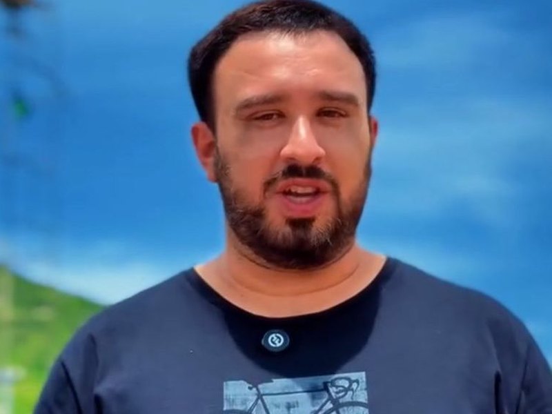 Antoniel Holanda de camiseta azul-marinho fala para a câmera em frente ao letreiro de Itaiçaba