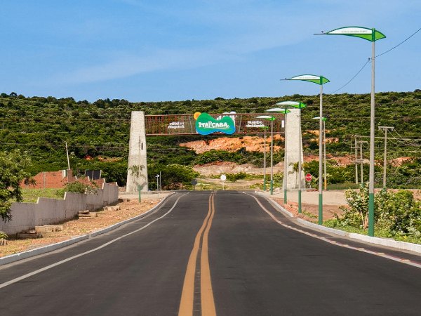 Avenida asfaltada com faixa amarela e postes leva ao pórtico de entrada da cidade